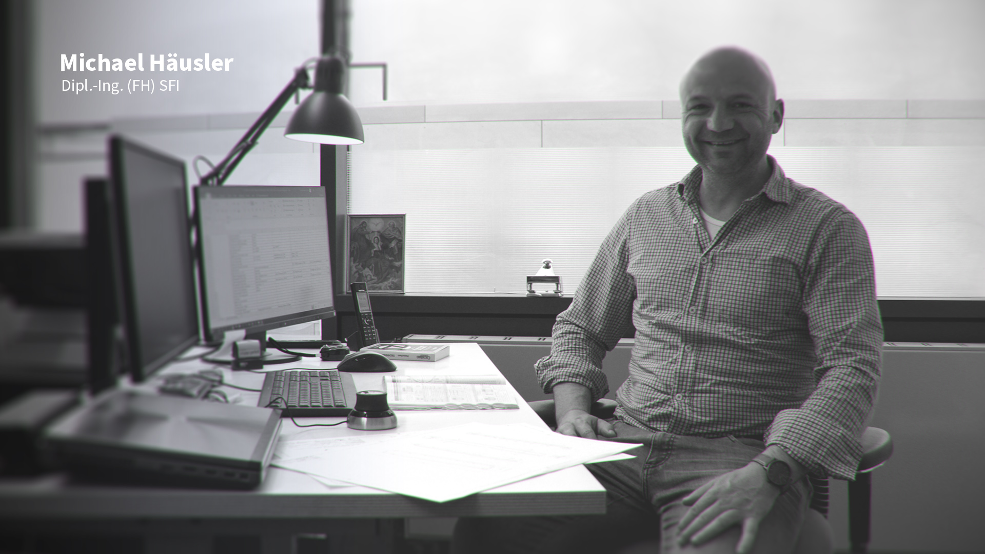 Mann in kariertem Hemd sitzt lächelnd an einem Schreibtisch mit zwei Monitoren, Tastatur, Telefon und Unterlagen in einem hellen Büro.